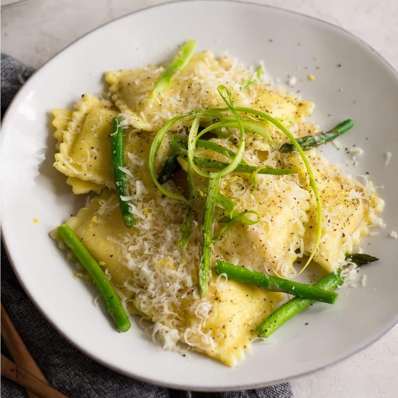 Ravioli pasta topped with grated cheese, black pepper, and fresh asparagus on a white plate.